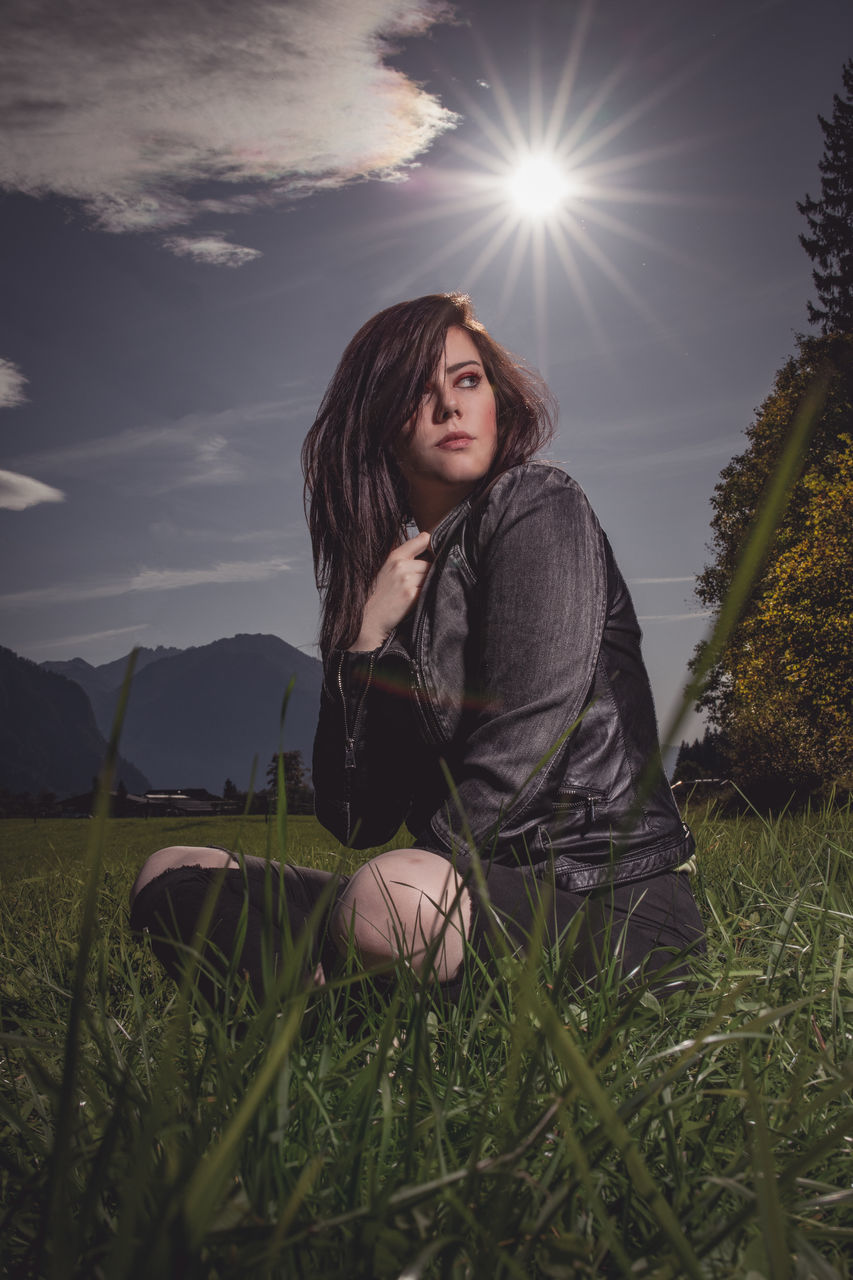 sky, field, land, one person, real people, young women, young adult, nature, leisure activity, lifestyles, plant, grass, beauty in nature, three quarter length, sunlight, sitting, adult, women, hairstyle, beautiful woman, outdoors, contemplation, lens flare