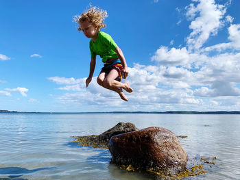 Boy jumping on rock in sea against sky