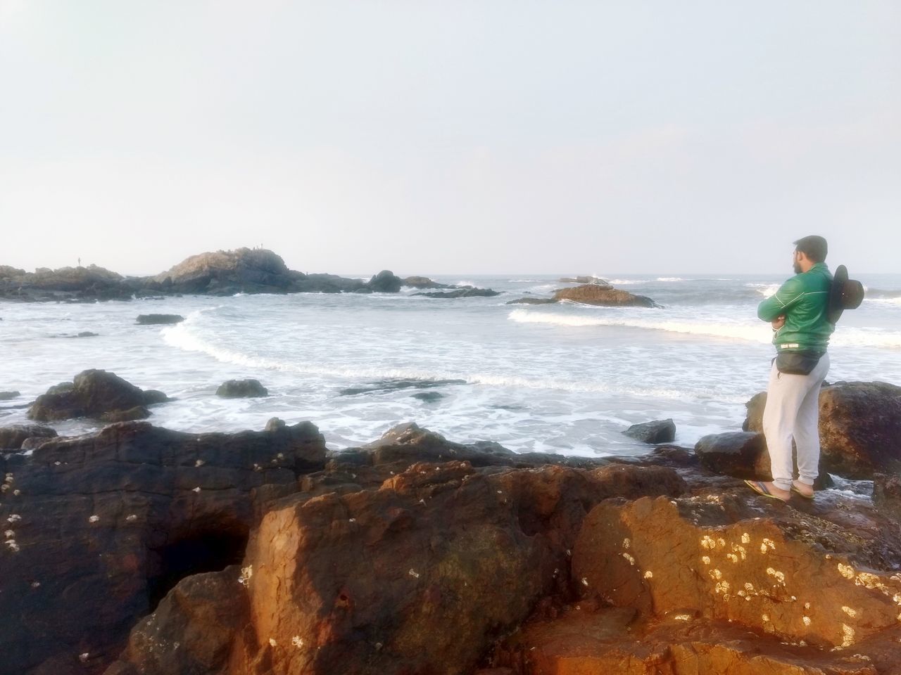 REAR VIEW OF MAN STANDING ON ROCK AT BEACH