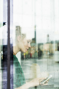 Young using smart phone while sitting on sill seen through glass at railroad station