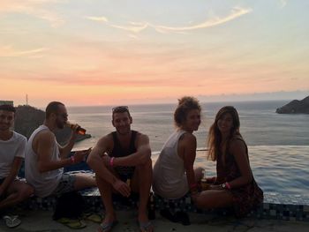 Friends sitting on beach against sky during sunset