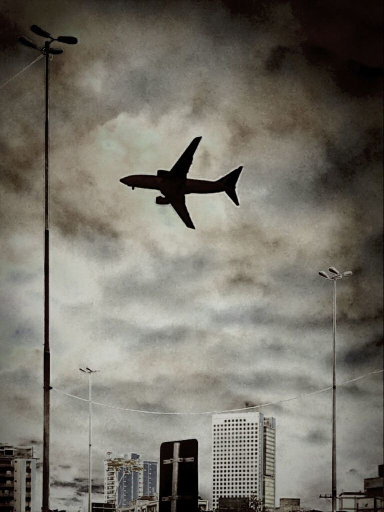 flying, airplane, sky, low angle view, air vehicle, cloud - sky, building exterior, mid-air, transportation, mode of transport, built structure, cloudy, architecture, street light, city, cloud, travel, on the move, silhouette, public transportation