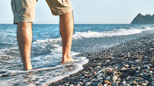 Male legs walking on pebble beach along the shore near the water with waves, low section.