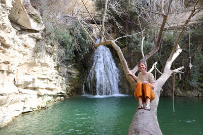 Smiling woman sitting on tree trunk against waterfall