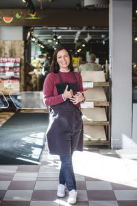 Full length portrait of smiling mature saleswoman wearing apron while standing in store
