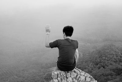 Rear view of man with hand raised sitting on rock against mountain