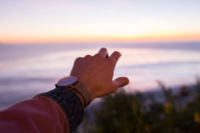 Cropped hand of man reaching sea against sky during sunset