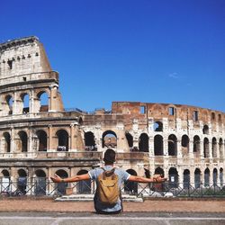 Rear view of man sitting with arms outstretched by colosseum against clear blue sky