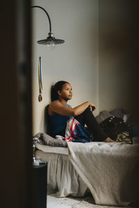 Teenage girl sitting on bed against wall in bedroom at home
