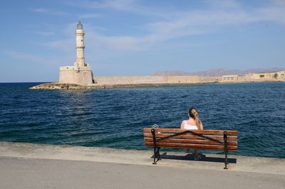 Rear view of woman sitting on bench against lighthouse in sea