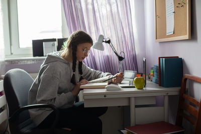 A teenage girl with glasses reads a book at her desk at home.