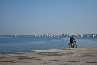 Man riding bicycle on city by sea against clear sky