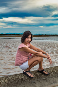 Portrait of mid adult woman crouching by sea against cloudy sky