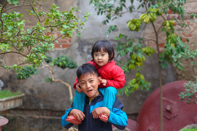 Happy smiling vietnamese boy holding his little sister sitting on his neck in the garden
