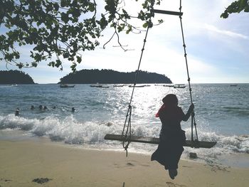 Rear view of woman swinging at beach against sky during sunny day