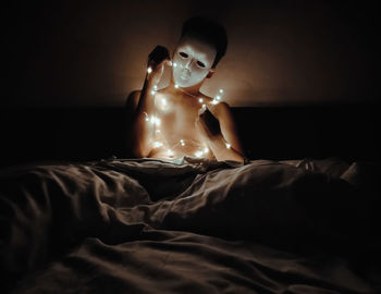 Shirtless young man with string lights wearing mask while sitting on bed in darkroom