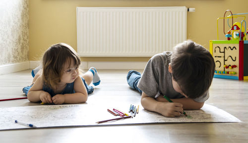 Siblings drawing in book while lying on floor at home