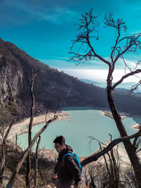 Young man standing by bare tree against lake and sky