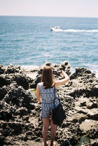 Rear view of young woman photographing with mobile phone while standing at beach