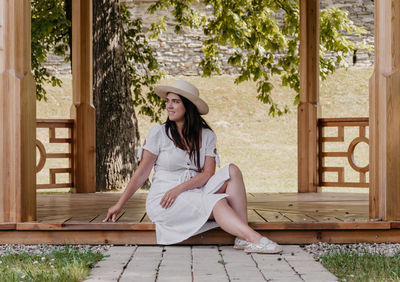 Portrait of beautiful young woman wearing white dress sitting under wooden pavilion in park.