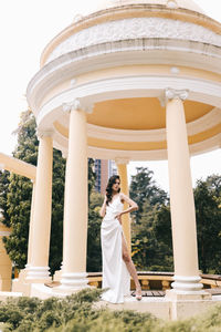 A beautiful brunette lady in an elegant wedding dress poses among the columns in the old city park