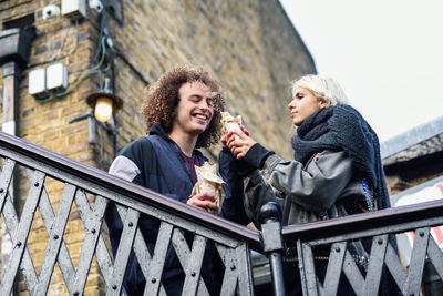 Low angle view of woman feeding boyfriend by railing against building in city