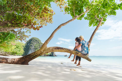Full length of mother and daughter sitting on tree trunk at beach