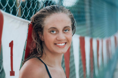 Portrait of smiling young woman by chainlink fence outdoors