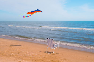 Kite flying over empty folding chair on shore at beach against sky