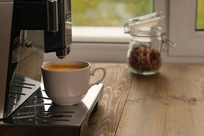 Automatic machine for coffee in white cup at window at daytime.  jar of hazelnuts on background