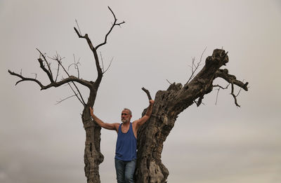 Portrait of adult man standing on bare olive tree trunk against sky
