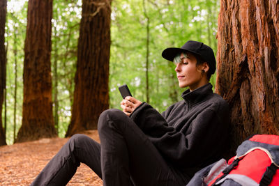 Side view of cheerful female tourist leaning on tree in woods and browsing smartphone while resting in monte cabezon natural monument of sequoias