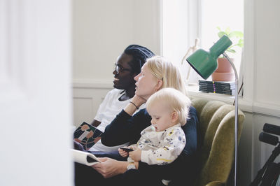 Side view of working mother with baby girl sitting by colleague on sofa in creative office