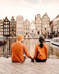 Rear view of couple holding hands while sitting by river against buildings in city