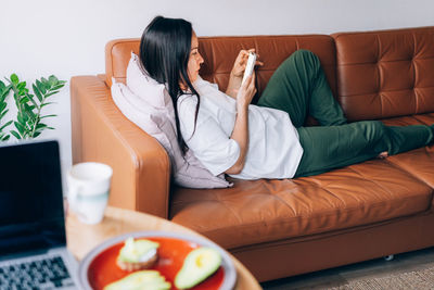 Portrait of young brunette female texting on smartphone while chilling on sofa at cozy apartment.