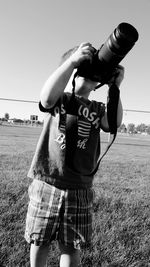 Boy photographing with camera while standing on field against clear sky