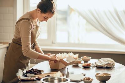 Chef in apron works in sunlit kitchen, fold, preparing box filled with pastries and other delights