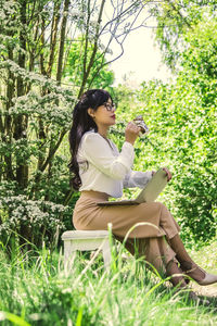 Side view of young woman using phone while sitting on plants