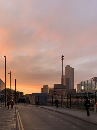 People on street by buildings against sky during sunset