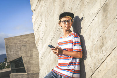 Boy holding phone while standing outside building on sunny day