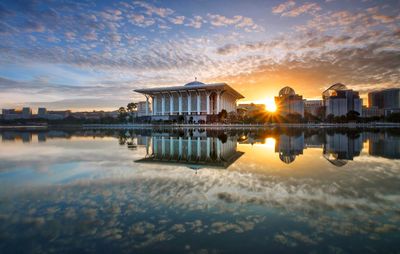 Buildings by lake against sky during sunset