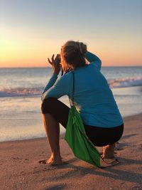 Woman crouching at beach against sky during sunset