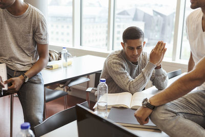 Serious student reading book while sitting with friends in classroom at university