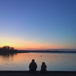 Silhouette couple sitting by lake against clear sky during sunset
