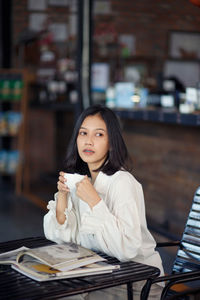 Young beautiful asian woman in white blazer with coffee cup sitting in cafe dark brown background