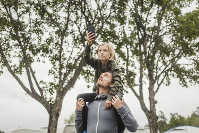 Girl holding mobile phone while sitting on father's shoulders against trees