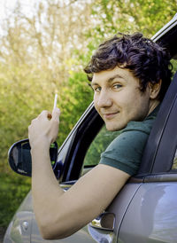 Portrait of young man looking through car window while holding smart phone
