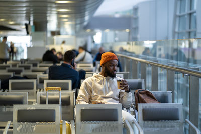 Young african man drinking coffee and eating sandwich while waiting for flying at airport terminal