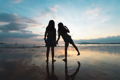 Silhouette girls holding hand standing on beach against sky