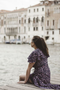 Woman sitting by canal against buildings in city